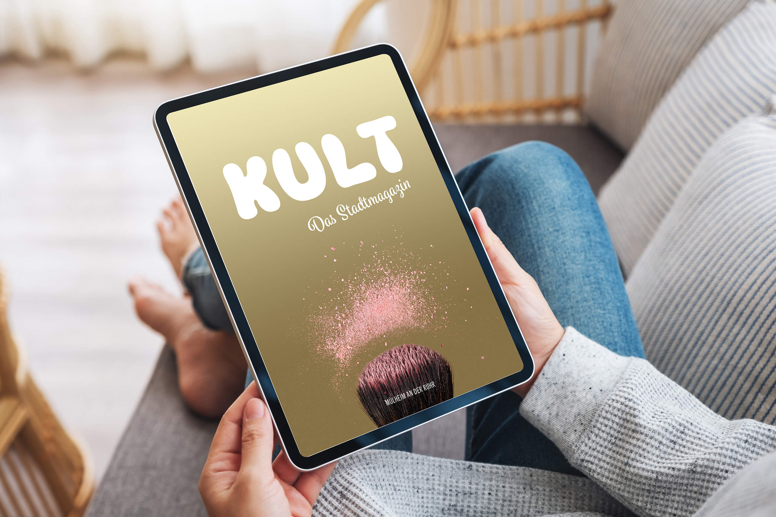 Frau auf einer Couch hält Tablet mit dem Titel einer Ausgabe von KULT – Das Stadtmagazin für Mülheim 