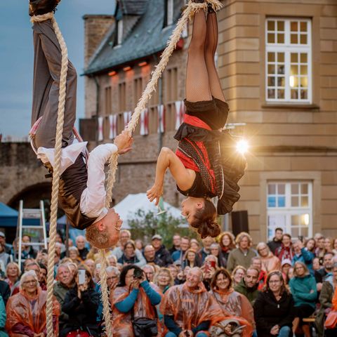 Broicher Schlossnacht 2025: Ein Artisten-Paar hängt mit den Füßen an Seilen und schenkt ein Glas Wasser ein, das Publikum sitzt im Innenhof von Schloß Broich und lacht begeistert.