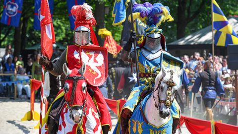 Ein rot und ein blau gekleideter Ritter mit Flaggen sitzen auf zwei Pferden mit passenden Gewändern; im Hintergrund ist die Kulisse des Ritterturniers und das Publikum zu erkennen.