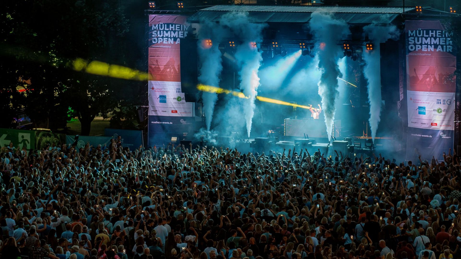 MÜLHEIM SUMMER OPEN AIR 2024: Das Publikum feiert vor der Bühne im MüGa-Park und Fontänen schießen Nebel in die Luft.
