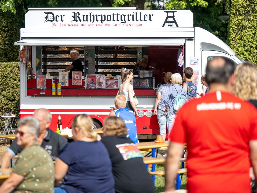 Gäste stehen am Stand von „Der Ruhrpottgriller”, wo Speisen zubereitet werden. Weitere Menschen sitzen au Bierbänken und genießen das sonnige Wetter.
