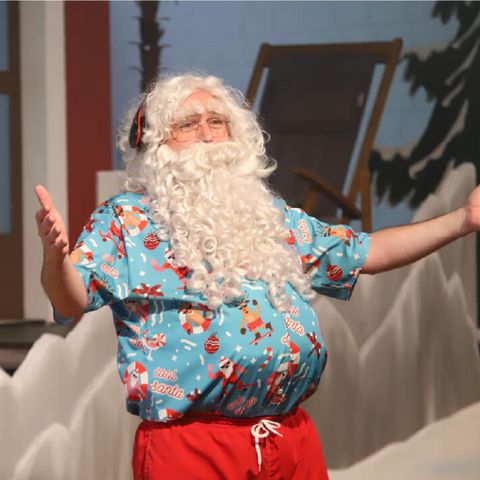 Verkleideter Santa Claus mit langem weißem Bart und Sommerhemd steht vor einer Bühnenkulisse und hebt feierlich die Arme.