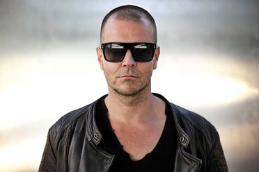 Imagefoto: MOGUAI trägt Sonnenbrille und Lederjacke, steht vor einem beleuchteten grauen Hintergrund und blickt lässig in die Kamera. 