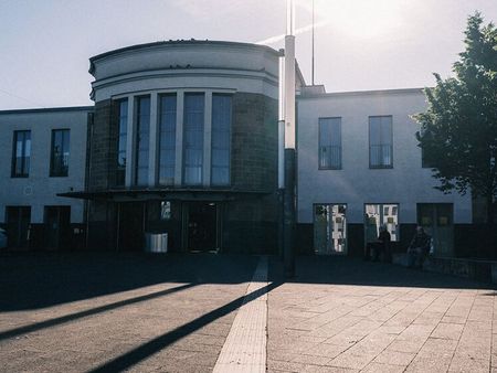 Asphaltierter Boden vor dem Eingang eines Bahnhofs, die Sonne strahlt über dem Gebäude 