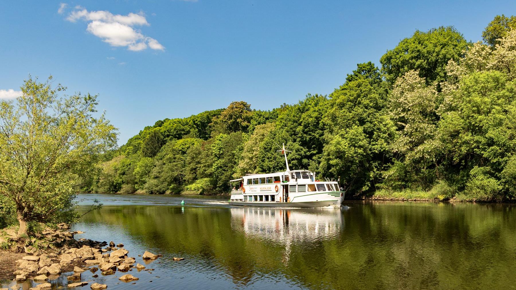 Weißes Schiff fährt bei sonnigem Wetter auf einem Fluss, umgeben von idyllischer Natur 