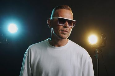 Imagefoto: C-Deluxe trägt Sonnenbrille und weißes Shirt und steht mit lässiger Körperhaltung vor einem dunklen Hintergrund mit einem blauen und gelben Scheinwerfer.