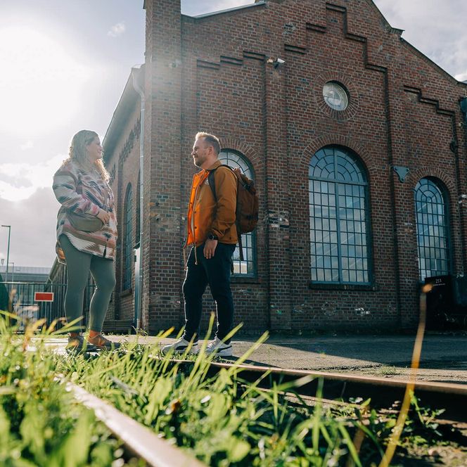 Zwei junge Erwachsene stehen an Bahnschienen, die mit Gras bewachsen sind. Im Hintergrund ragt eine Dreherei mit großen Fenstern in den Himmel.