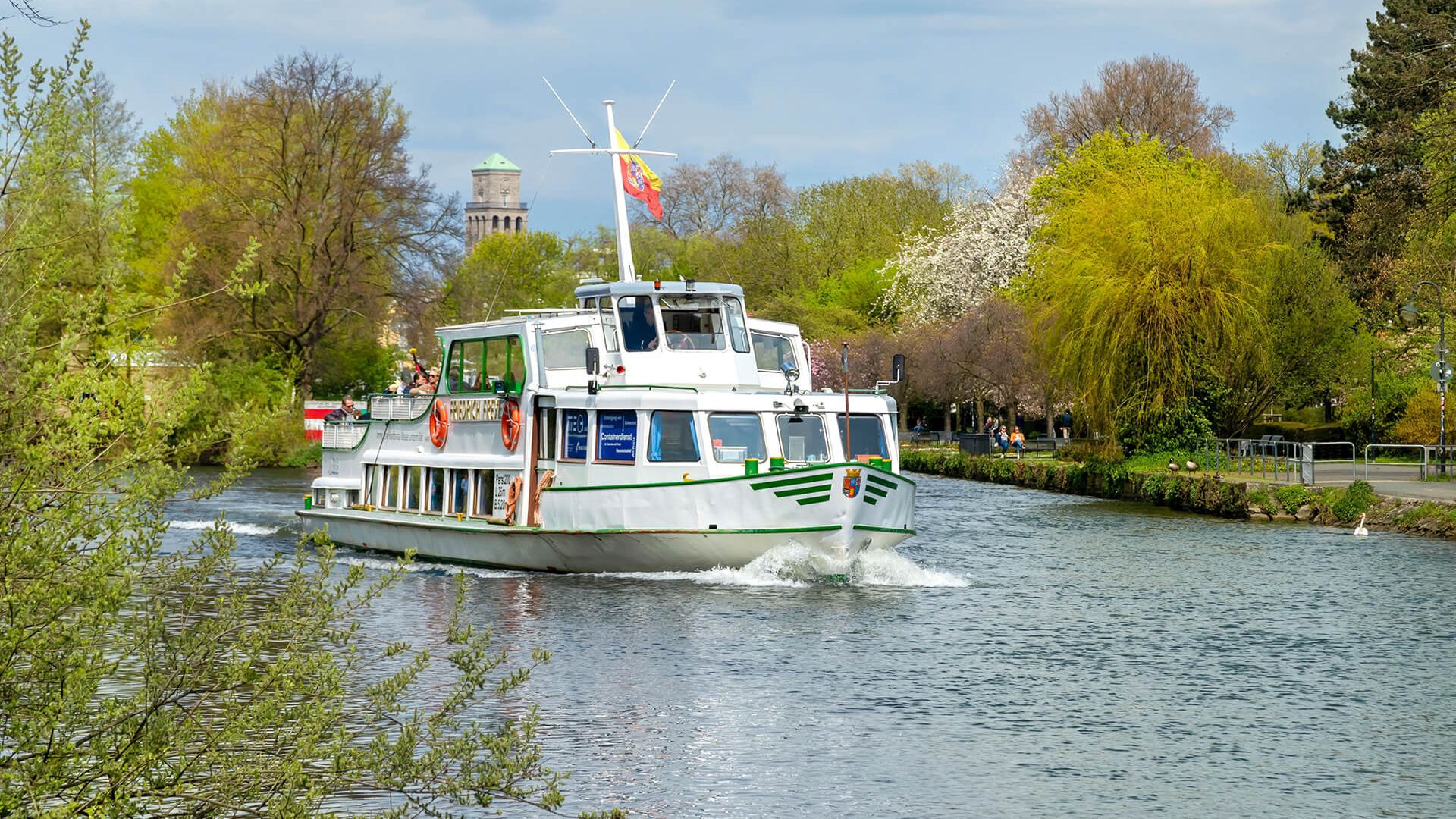 Ein Schiff der Weißen Flotte fährt über die Ruhr, das Ufer ist gesäumt von grünen Pflanzen, im Hintergrund erkennt man den Rathausturm der Stadt Mülheim an der Ruhr