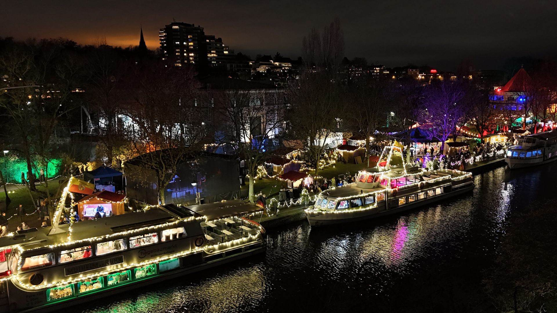 Schiffe mit Beleuchtung am Ufer der Schleuseninsel, wo Gäste auf der Schiffsweihnacht über den beleuchteten Weihnanchtsmarkt bummeln.