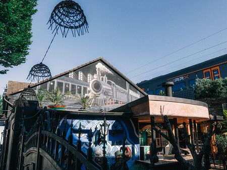 Viele bunte Kunstwerke zieren ein Haus mit einer Open-Air-Bar, oberhalb fährt ein Zug vorbei