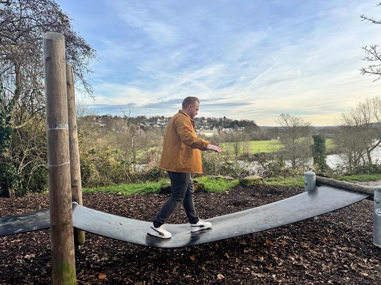 Junger Mann balanciert auf einer Hängematte aus Gummi. Im Hintergrund sieht man viel Natur, einen Fluss und das Farbenspiel des Himmels.