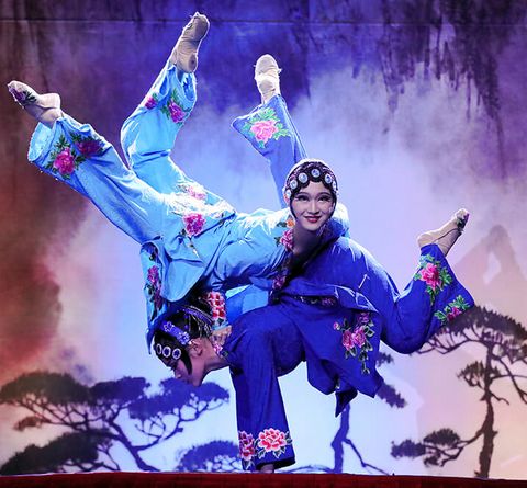 Zwei Frauen in blauen Köstum zeigen gymnastische Pose vor einer rot-violetten Kulisse mit Nebelwolken