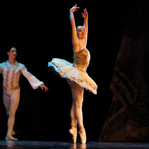 Balletttänzerin in hellem Tutu tanzt auf Spitze auf einer dunklen Bühne; im Hintergrund steht ein Tänzer.