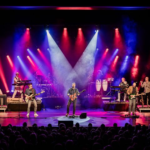 Band spielt vor Publikum auf einer Bühne; Scheinwerfer in rot und violett von oben herab