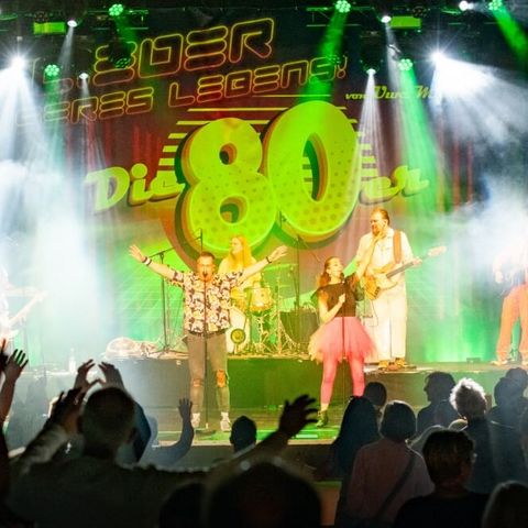 Show „Lieder unseres Lebens – Die 80er”: Musiker und Musikerinnen steht auf einer Bühne und sorgt für Stimmung im feiernden Publikum.