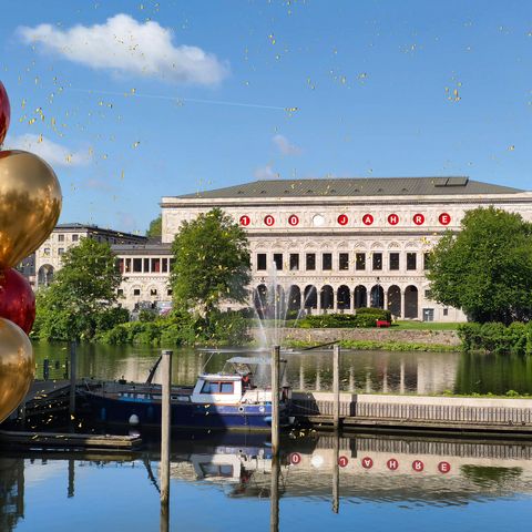 Blick über die Ruhr auf die Stadthalle Mülheim mit Schriftzug „100 Jahre” bei sonnigem Wetter, umgeben von roten und goldenen Ballons (retuschiert)