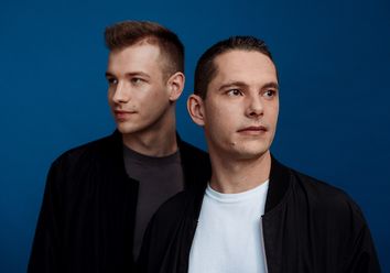 Imagefoto: Das Duo Gestört aber GeiL steht in schwarzer und weißer Kleidung vor einem dunkelblauen Hintergrund und blickt entspannt in entgegengesetzte Richtungen.