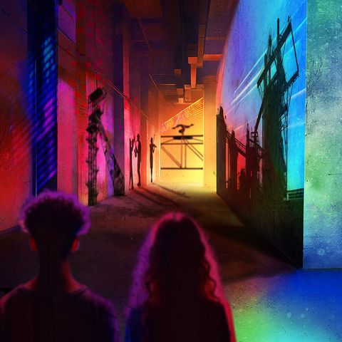 Zwei Menschen laufen einen langen bunt beleuchteten Gang entlang, an dessen Wänden Schatten von Sehenswürdigkeiten im Ruhrgebiet zu erkennen sind, am Ende des Gangs führt eine Akrobatin ein Kunststück auf.