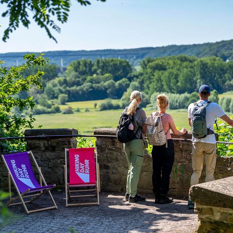 Drei Wandernde stehen auf einer Plattform und genießen den Ausblick auf das Mülheimer Ruhrtal, links stehen zwei Liegestühle mit der Aufschrift „Hiking Day Ruhr” 